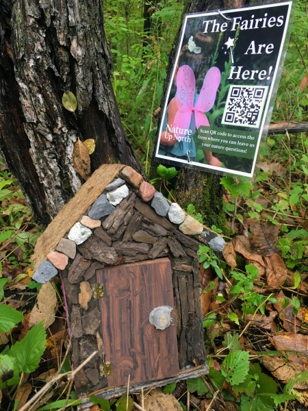 Image of a brown fairy house at the base of trees with a poster attached to a tree