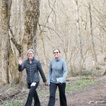 A woman in dark grey sweater and women in light gray sweater walking down a wooded trail and waving at the camera