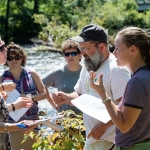 5 people standing in a circle in front of a river all holding pieces of paper and looking at a clear vial of water