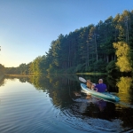 Sun setting over a glassy smooth dark blue river with dark green pine trees and someone paddling a kayak