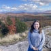 Women with brown hair and light blue sweater on a rock in front of landscape of green, orange, and red fall foliage