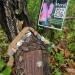 Image of a brown fairy house at the base of trees with a poster attached to a tree